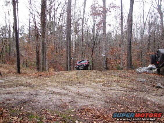 red Jeep Wrangler driving up a dirt hill in a wooded area, another Jeep is parked to the right