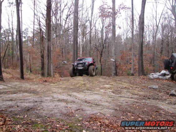 Red Jeep Wrangler on a dirt mound in a wooded area, with another dark SUV visible to the right