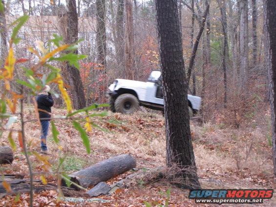 White off-road vehicle with large tires climbing a wooded hill. Person stands to the left