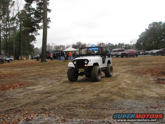 White Jeep CJ with big tires driving on dirt, people and tents in background