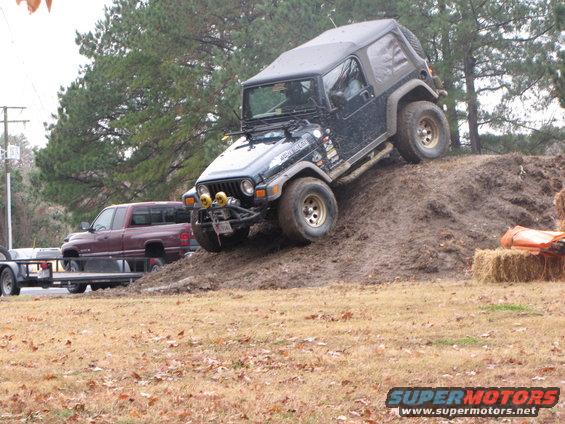 Black Jeep Wrangler climbing a dirt hill, with a red pickup truck and trailer in the background