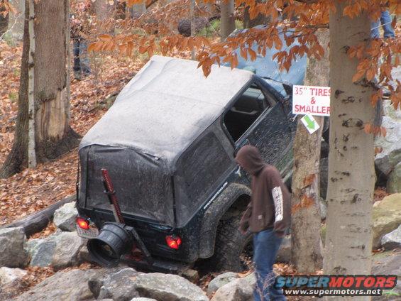 Off-road vehicle with soft top stuck on rocks, person in hoodie walks by. Sign says '35" TIRES & SMALLER'