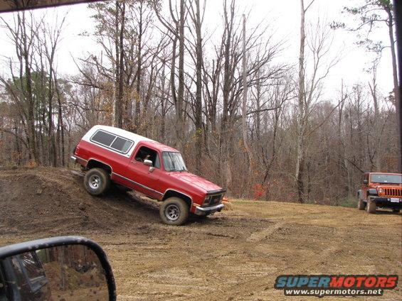 Red SUV with white camper shell driving up a dirt hill, orange Jeep in background