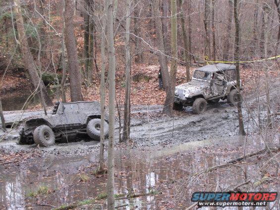 Two modified off-road vehicles covered in mud, one partially submerged in a muddy ditch, the other on a muddy trail in a wooded area