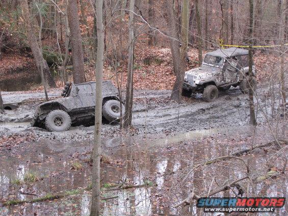 Two muddy off-road vehicles navigating a wooded trail with deep mud and water