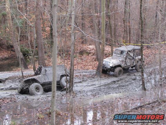 Mud-covered Jeep Wrangler and another off-road vehicle partially submerged in a muddy forest trail