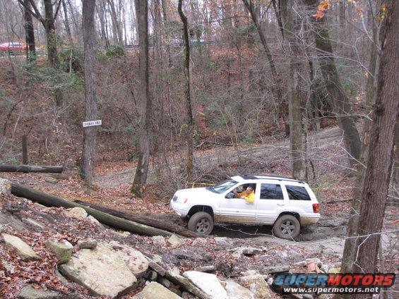White Jeep Grand Cherokee navigating a muddy, wooded trail with fallen logs