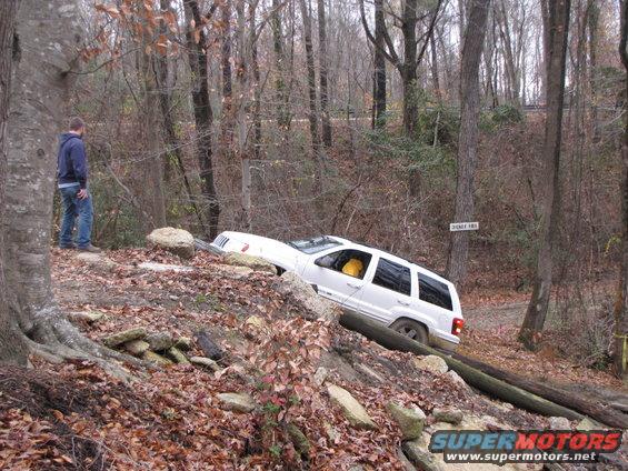 White SUV tilted on a wooded trail with a person watching