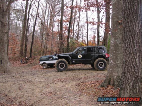 Black modified SUV with large tires parked next to a white and black SUV in a dirt clearing surrounded by trees