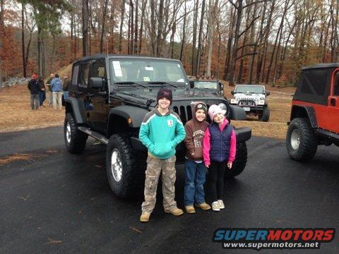 Black Jeep Wrangler with three children standing in front. Other Jeeps visible