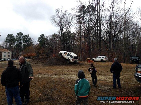 White SUV on a dirt mound with people watching. Another SUV and a Jeep are parked nearby