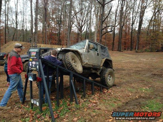 Silver Jeep Wrangler on a metal ramp, with two men nearby. Muddy terrain and trees in background