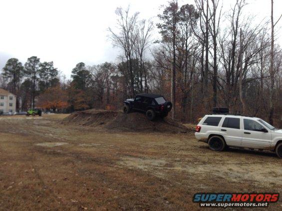 black SUV climbing dirt mound, white SUV parked nearby, green Jeep in background