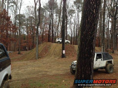 Off-road course with a white Jeep and two white SUVs on a dirt hill. Trees and fallen leaves surround the area