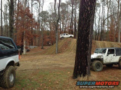 Off-road vehicles on a dirt track in the woods. One white Jeep is in the foreground, and a white SUV is on a hill in the background. A black and white SUV is partially visible on the left