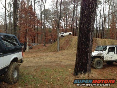 Black and white Ford Bronco side view, muddy tires, off-road scene with trees and dirt mounds