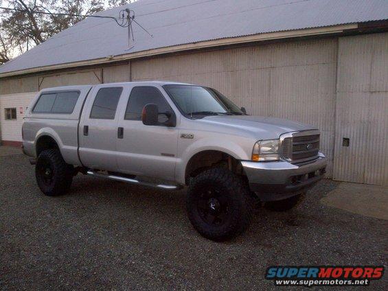 Silver Ford F-250 Super Duty pickup truck with a camper shell and lifted suspension, parked in front of a corrugated metal building