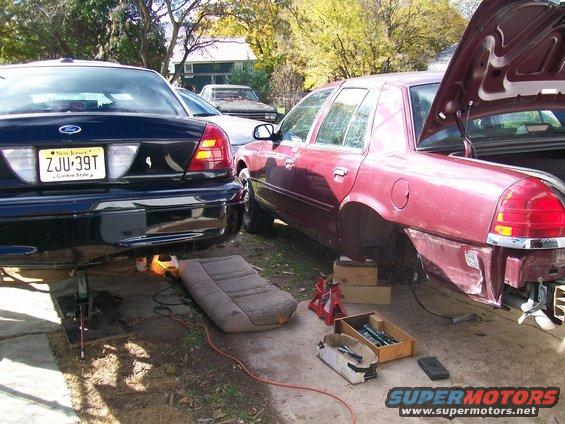 Two Ford Crown Victorias, one black and one maroon, are parked outside with their rear ends raised on jacks. The maroon car is missing its rear bumper and has its trunk open