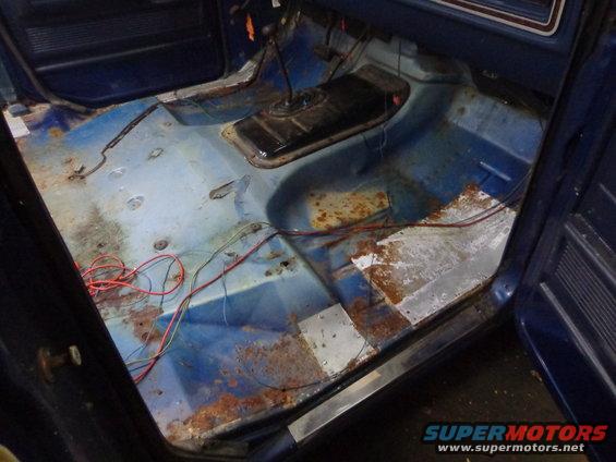 Interior of a blue vehicle showing rusted floorboards and wiring