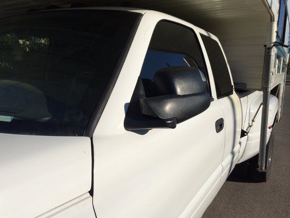 Side view of a white pickup truck, possibly a Dodge Ram from the late 90s. It has large side mirrors and a camper shell in the bed