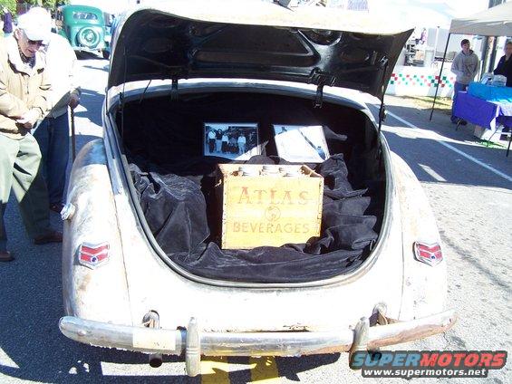 Trunk open showing a wooden crate labeled 'ATLAS BEVERAGES', two framed photos, and black fabric lining. Another car is visible in the background