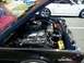 1971 Ford Ranchero - The engine bay of a black classic car is open, revealing its mechanical components. A modern silver convertible is parked behind it.