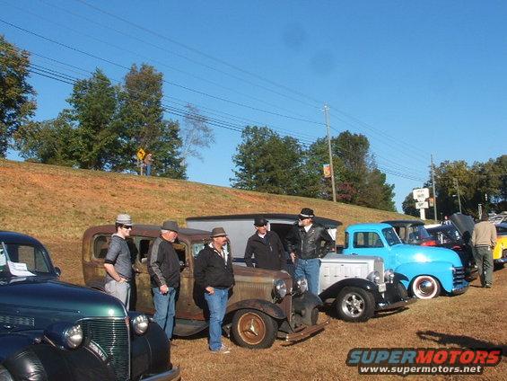 Several old cars parked outside with men in hats standing around them