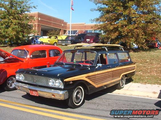 Black Ford Country Sedan wagon with wood paneling and surfboards on roof rack. Red interior visible. Parked next to a red car