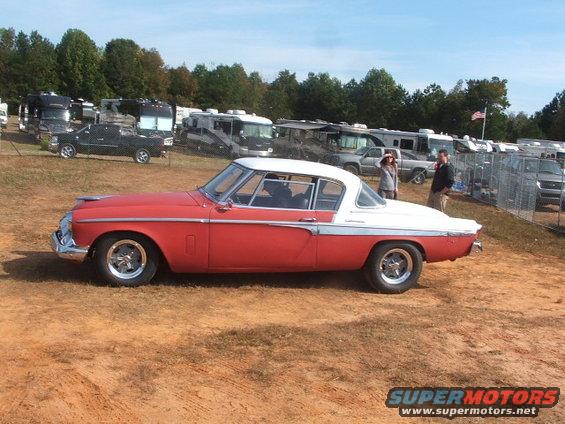 Red and white Studebaker Hawk coupe parked on dirt lot with RVs and other vehicles in background