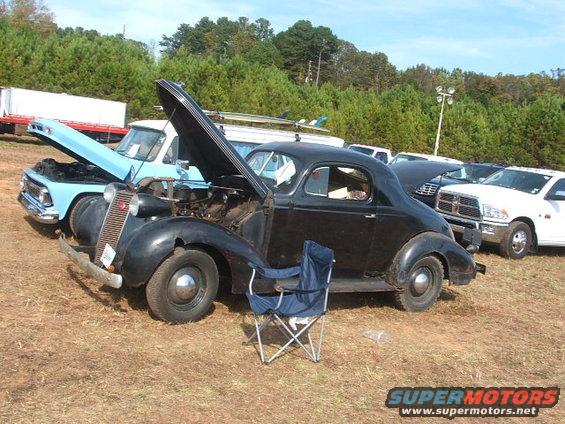 Vintage black coupe with hood open, parked next to a blue pickup truck and other vehicles