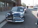 1971 Ford Ranchero - A classic dark gray car from the 1950s is parked on the street, with other vehicles and people in the background.