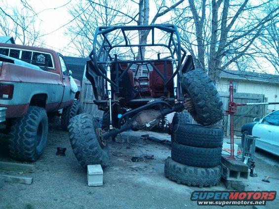 Off-road buggy frame with roll cage, large tires stacked, and a red pickup truck nearby