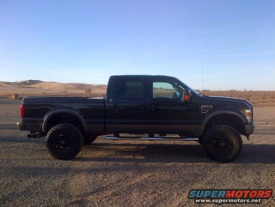 Black crew cab pickup truck with aftermarket wheels and tires, parked on a gravel surface under a clear blue sky