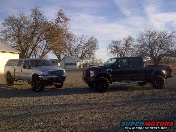 Two lifted pickup trucks, one silver with a cap, one black, parked on gravel