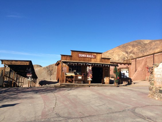 Wooden town hall building with a sign above the entrance. Another covered structure with signs is to the left. Desert landscape in background