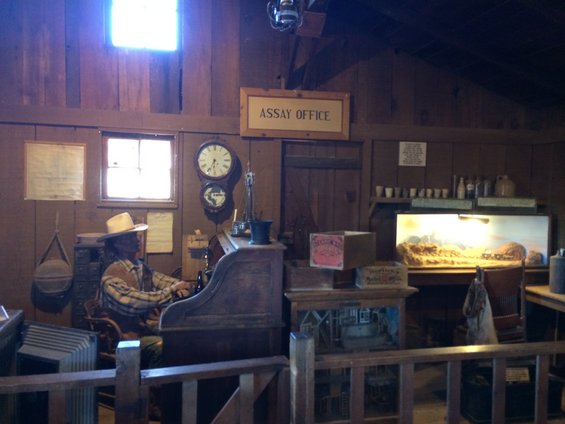 Mannequin dressed as prospector sits at desk in assay office with clocks and bottles on shelves