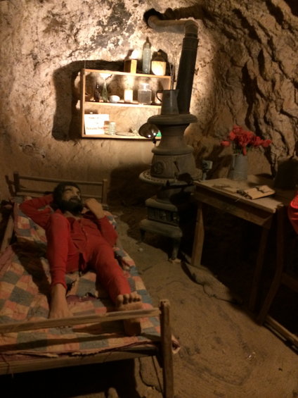 Man in red pajamas on a cot inside a cave with a wood stove and shelves