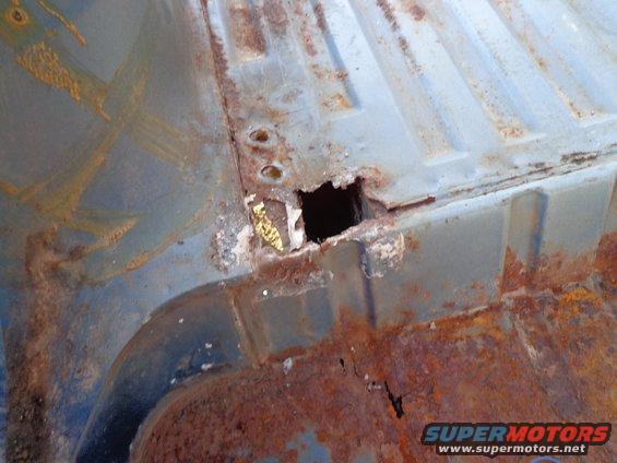 Rusted metal floor panel with a square hole, showing extensive corrosion and blue paint