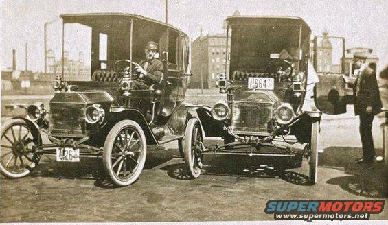 Two old cars parked side-by-side. One has a driver. A man stands by the other
