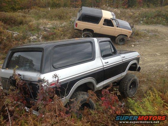 Two Ford Broncos, one silver and black, the other tan and black, are parked on a dirt trail with foliage around them