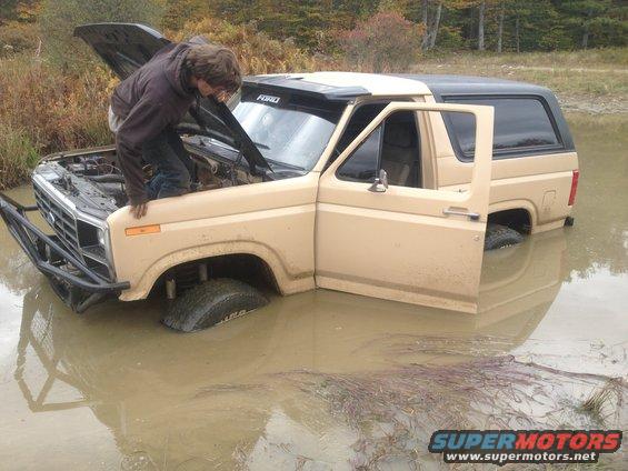 Ford Bronco stuck in muddy water, hood open, person looking at engine