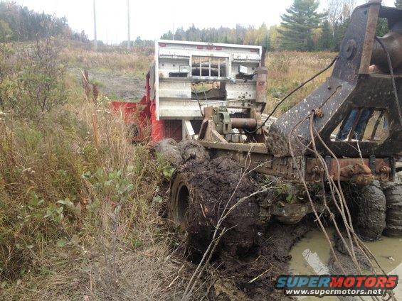 Mud-covered tow truck and trailer stuck in a ditch with tall grass and trees in the background