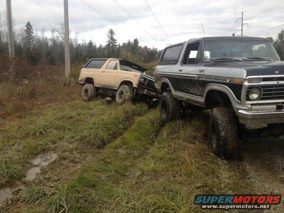 Two Ford Broncos stuck in muddy terrain, one tan and one two-tone black and silver