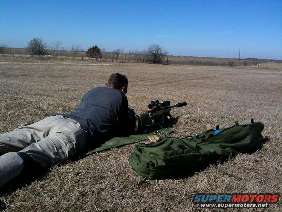 Man lying prone aiming a sniper rifle at a distant target on a dry grassy field under a clear blue sky