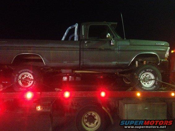 Side view of a dark green Ford pickup truck being towed on a flatbed trailer at night. It has a roll bar in the bed