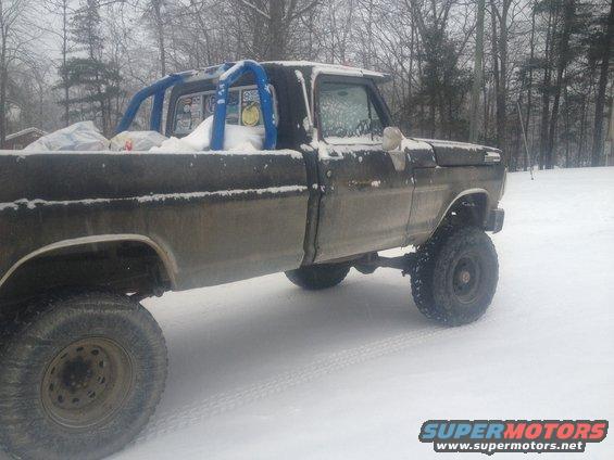 Dark-colored Ford F-250 pickup truck with a blue roll cage, lifted suspension, and snow on its body, seen from the side in a snowy environment