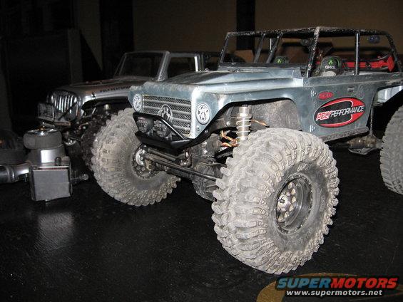 Two RC rock crawlers with large tires sit on a dark surface. One has a grille with a star emblem