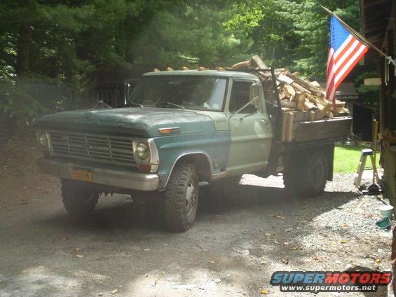 Green Ford F-Series truck loaded with firewood, parked outside a building with an American flag