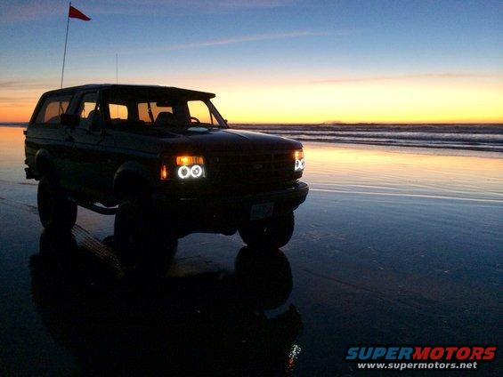 1992 Ford Bronco on wet sand with ocean waves and sunset in background