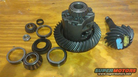 Differential gears and bearings laid out on a wooden surface. A differential case with a ring gear is central, with a pinion gear to the right. Several bearings and a nut are scattered around